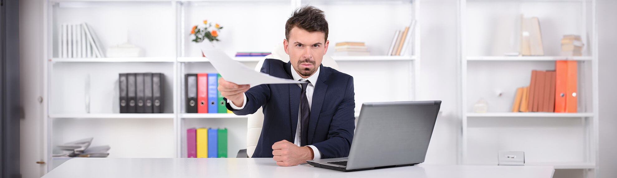 Mann im Anzug sitzt an einem Schreibtisch vor einem Laptop und hält mit ernstem Blick ein Dokument nach vorne.